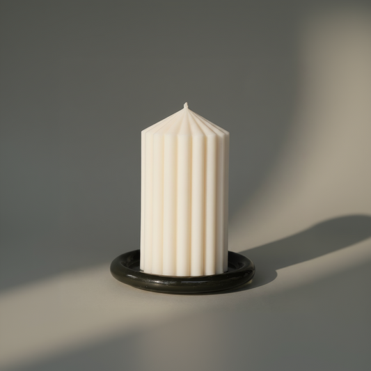 An off-white ribbed pillar candle standing upright in a glossy black circular holder against a neutral gray background.